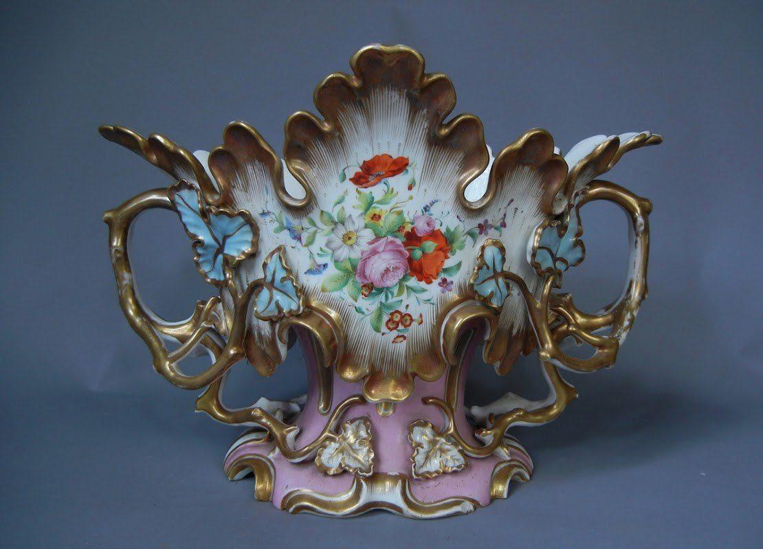 An ornate ceramic vase with scalloped edges, featuring floral designs in red, pink, and blue. The vase has intricate golden handles and detailing, set against a muted blue background.