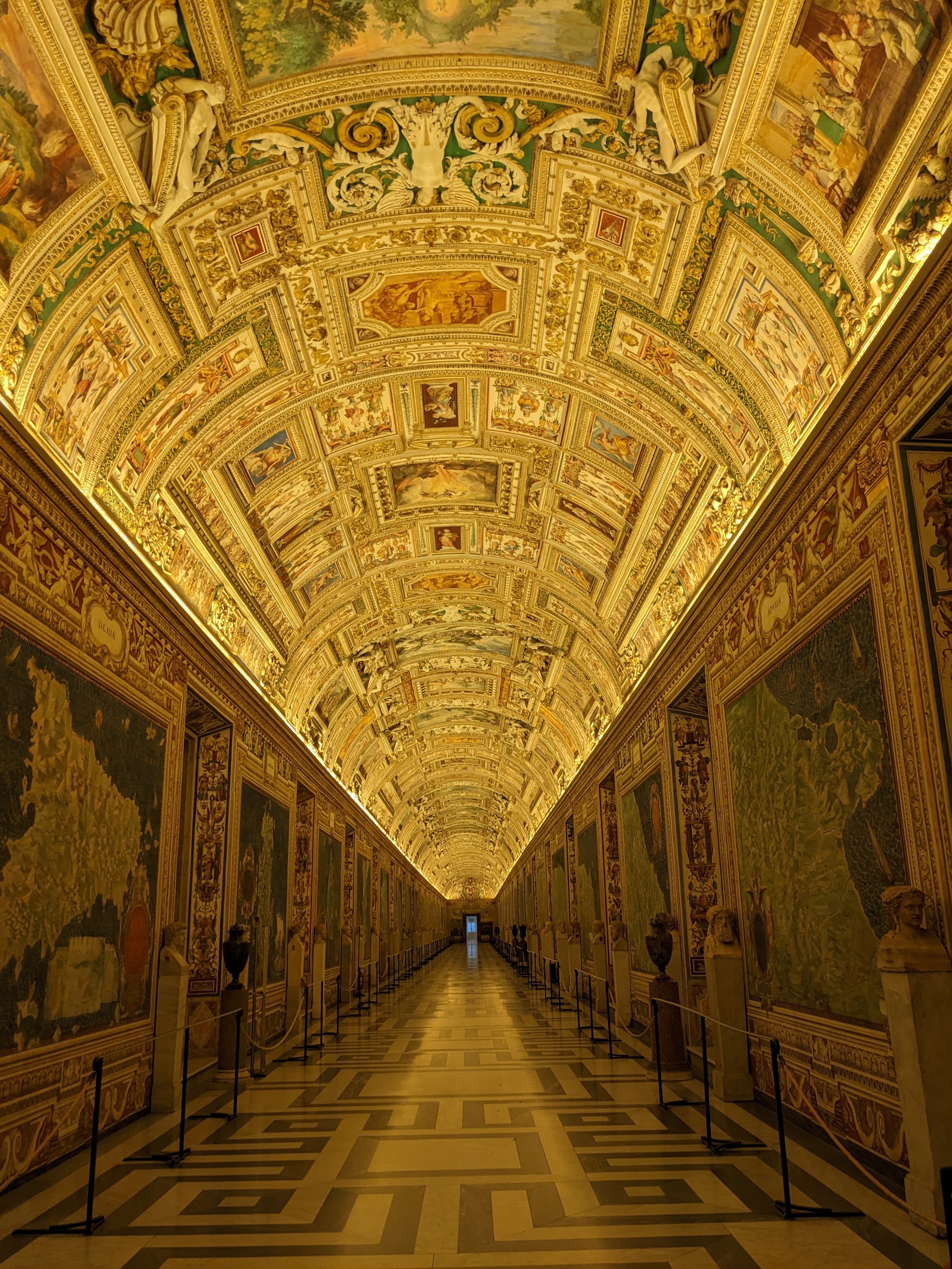 A grand, ornately decorated hallway with an arched ceiling covered in intricate frescoes and golden accents. The walls are adorned with detailed maps and artwork, and the floor features a geometric pattern. The hallway extends into the distance.