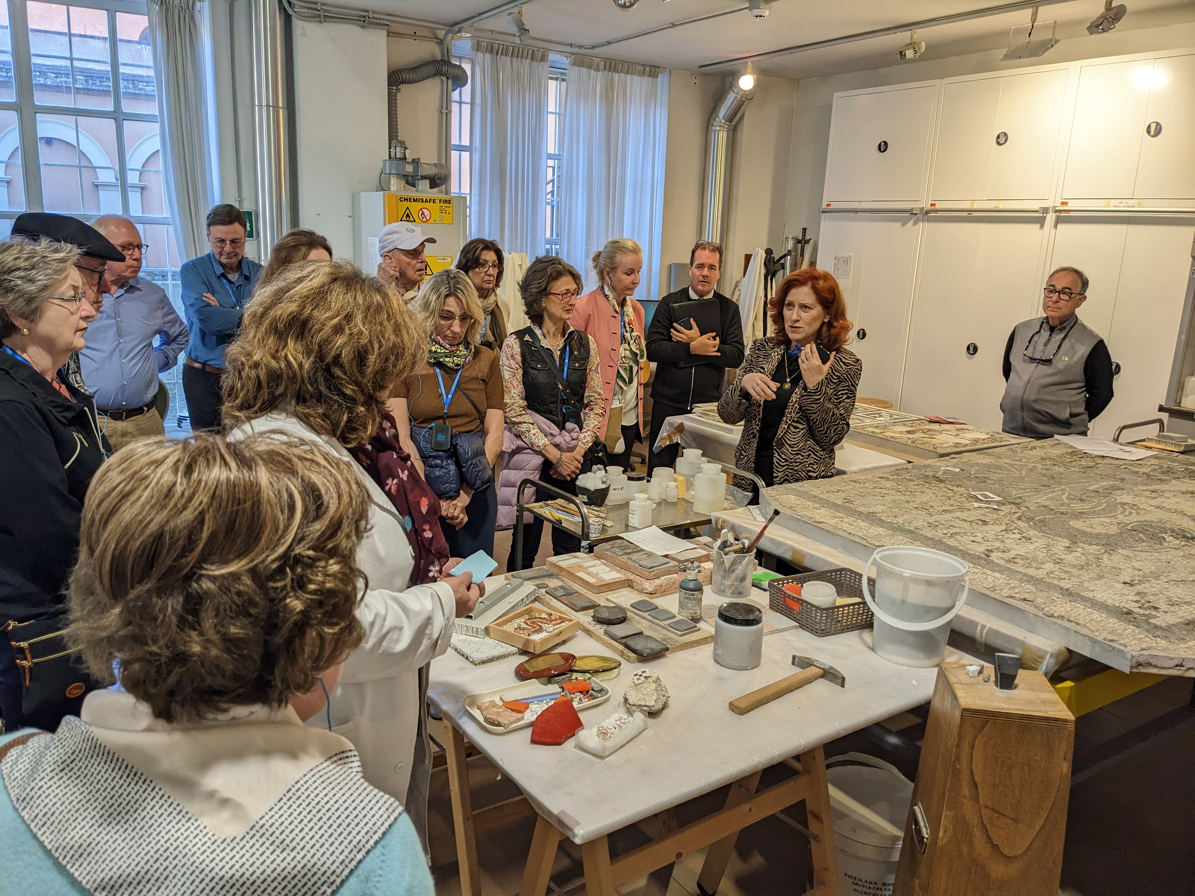 A group of people attentively listen to a woman explaining in a room filled with art restoration tools and materials. A large piece of artwork is placed on a table. The setting is well-lit with curtains and cabinets in the background.