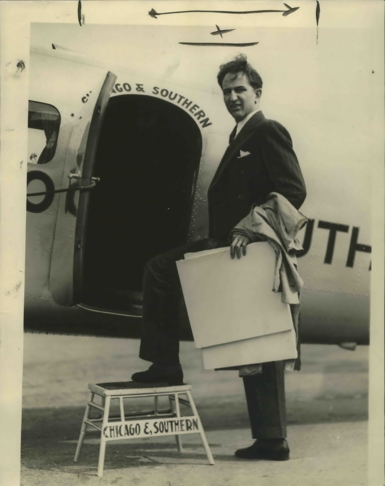 Man in a suit holding a coat under his arm, stepping onto a small stool labeled Chicago & Southern to board an airplane. The planes door is open, displaying part of the airlines name.