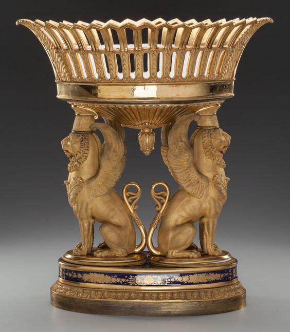 A lavish, gold centerpiece featuring a woven basket top supported by four ornate winged lion figures. The base is decorated with intricate patterns, creating a regal and artistic design.
