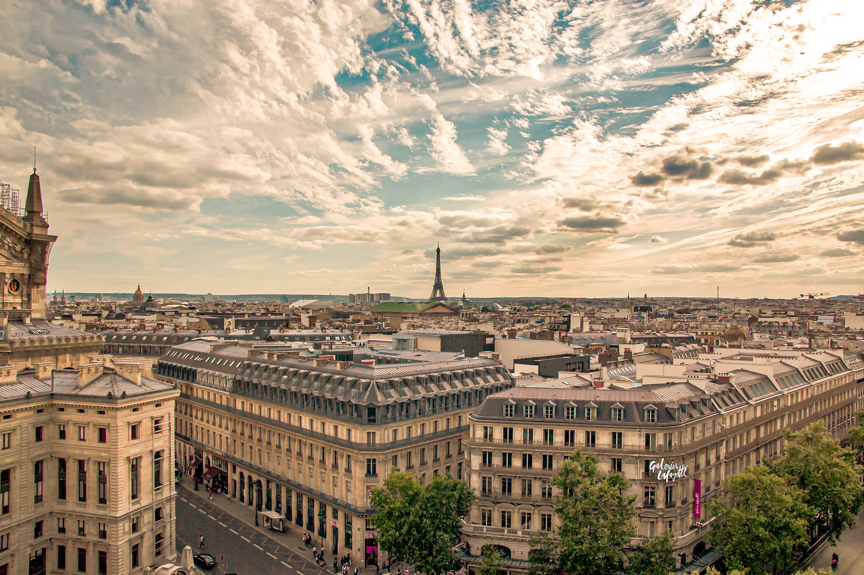 Aerial view of Paris, featuring historic buildings and the Eiffel Tower in the distance under a partly cloudy sky. The scene is bathed in warm sunlight, highlighting the architectural details and creating a vibrant cityscape.