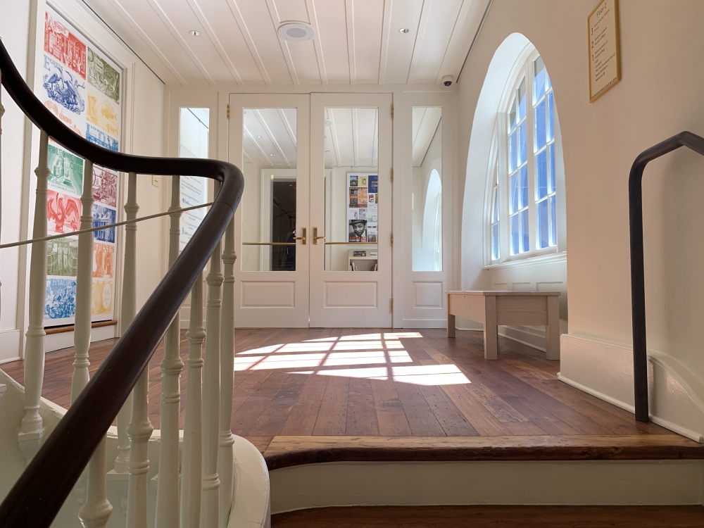 A bright, airy room with wooden floors and a spiral staircase. Large arched windows on the right let in natural light. On the back wall, colorful artwork and a glass door leading to another room. A bench sits below the windows.