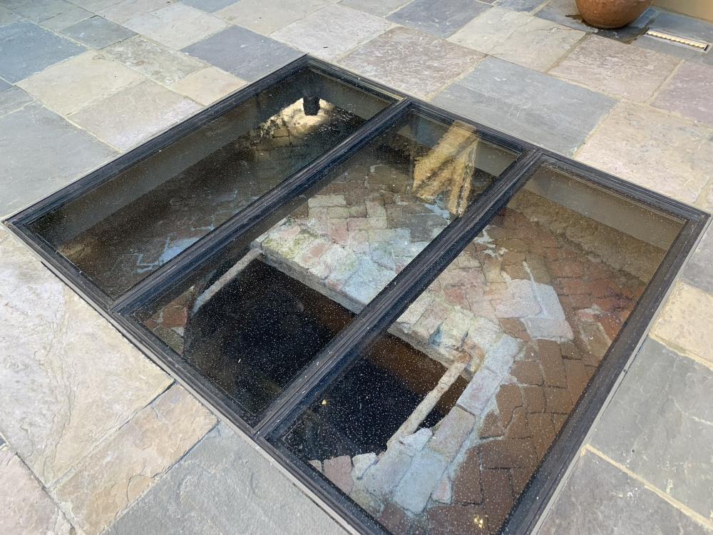 A glass-covered archaeological site is set within a stone-tiled floor, showcasing an ancient brick structure partially underground.