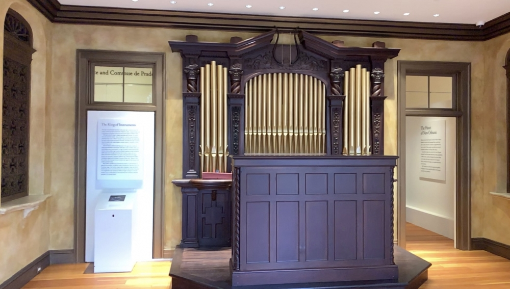 A historic church organ with ornate wooden casing and visible golden pipes. The room has wooden floors and decorative walls with informational panels on either side.