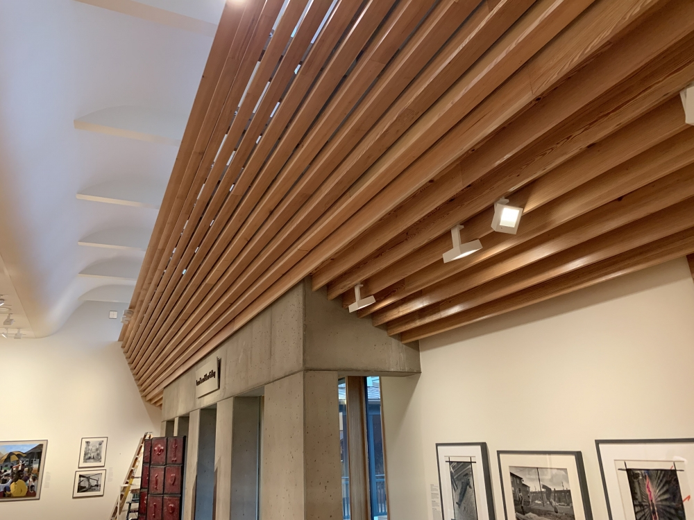 Interior view of an art gallery with a modern wooden slatted ceiling, concrete walls, and framed artwork on the walls. A ladder is visible in the corner, and track lighting illuminates the space.