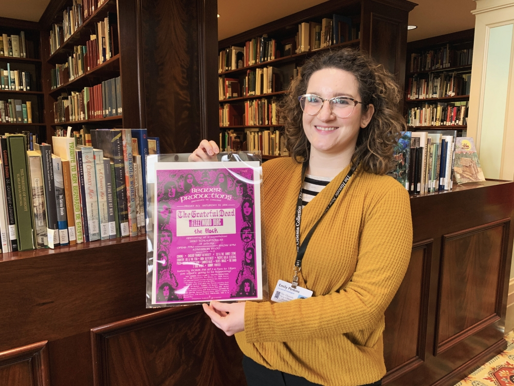 A person with curly hair and glasses is smiling and holding a framed poster in a library. The poster has a purple decorative border. Bookshelves filled with various books are visible in the background.