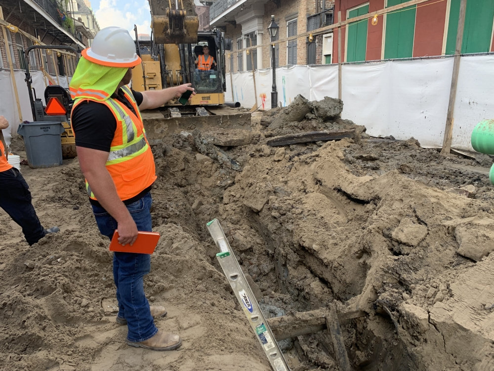 A construction worker wearing a hard hat and safety vest points at an excavation site. Behind him, another worker operates a large excavator. The area is surrounded by buildings, with a partially dug trench in the foreground.