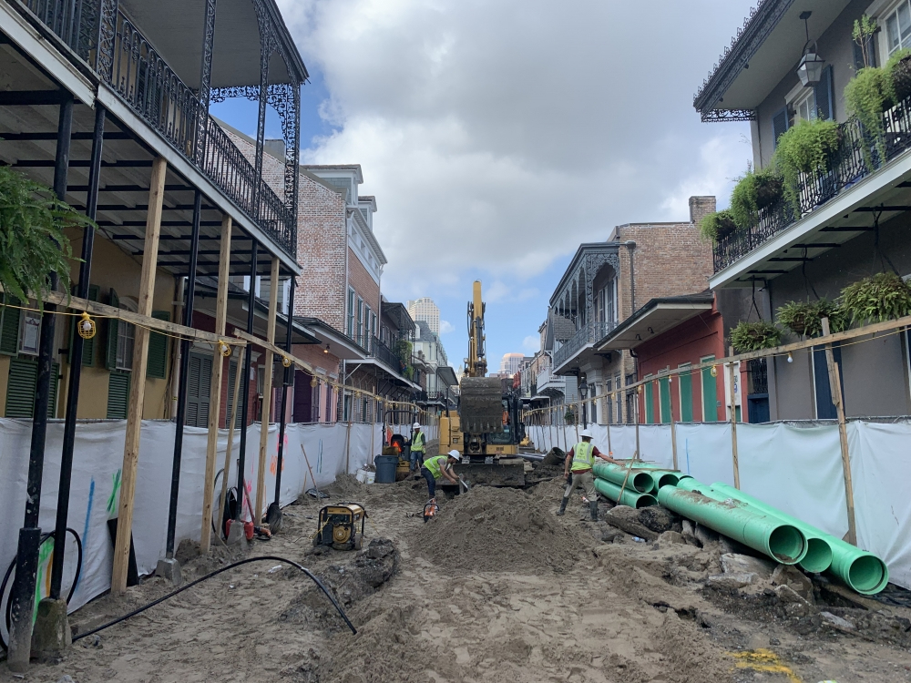 Construction workers operate machinery on a city street under renovation. Green pipes are lined up on the ground, and buildings with balconies border the street. The sky is partly cloudy.