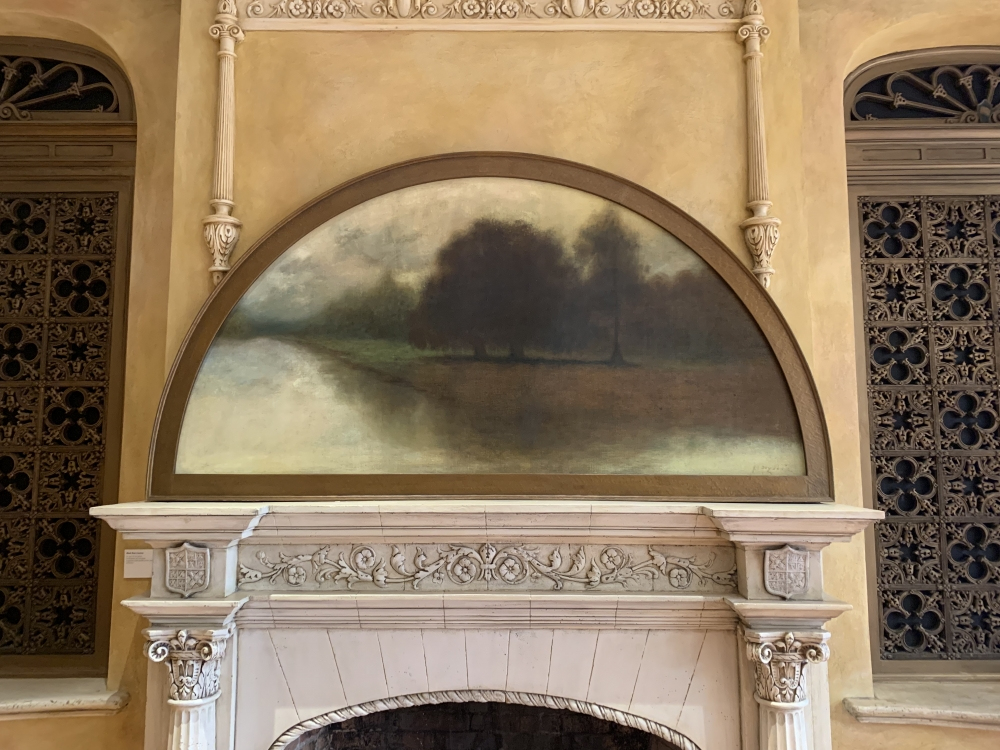 An elegant, antique fireplace mantle with ornate carvings set against a textured, yellowish-brown wall. Above the mantle is a semi-circular painting depicting a serene landscape with trees and a reflective body of water.