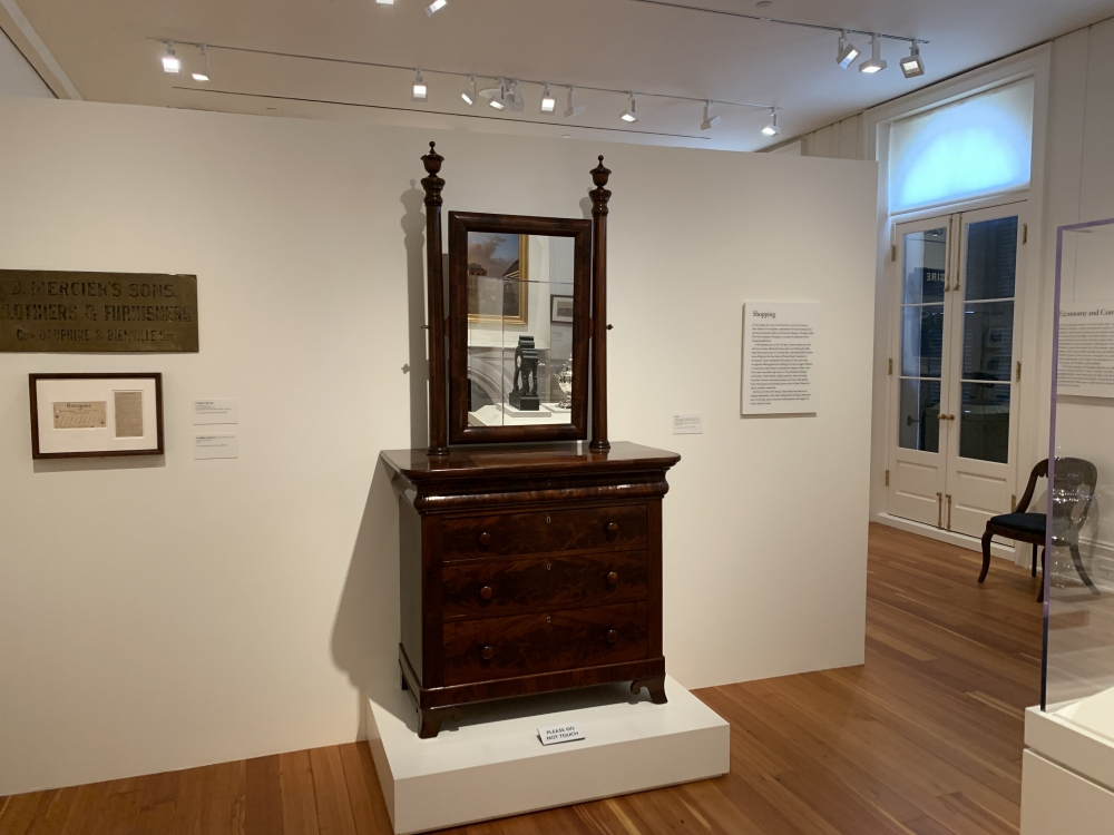 A wooden dresser with a mirror is displayed in a museum gallery. The dresser has four drawers and ornate features. Nearby, historical plaques and documents are mounted on white walls. The room has wooden floors and ample lighting.