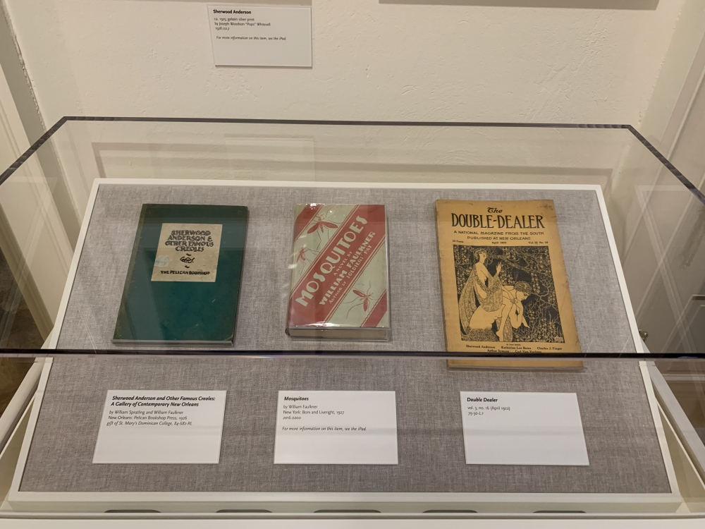Three vintage books displayed under a glass case on a gray fabric surface. They include Sherwood Anderson and Other Famous Creoles, Motu Mode, and The Double Dealer, each with descriptive labels below them.