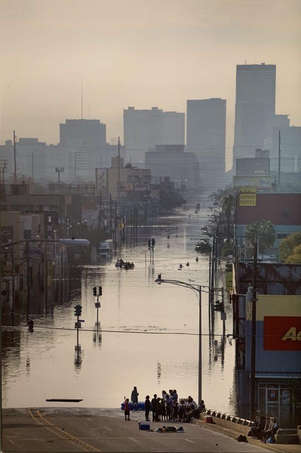 A city street is heavily flooded, with water reaching building heights. People gather on a section of dry road in the foreground. The skyline of a city with high-rise buildings is visible in the background, partially obscured by mist.