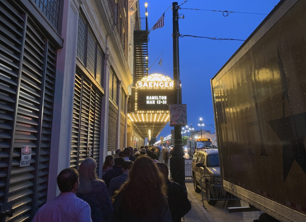 People stand in line outside the Saenger Theater at dusk. The marquee advertises a showing of Hamilton from March 12-31. A large trailer is parked nearby, and city lights illuminate the street.