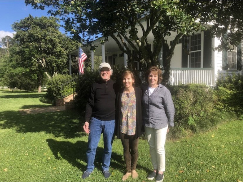 Three people stand smiling in front of a white house with a porch, shaded by trees. An American flag is displayed in the garden. They are casually dressed, enjoying a sunny day on a well-kept lawn.
