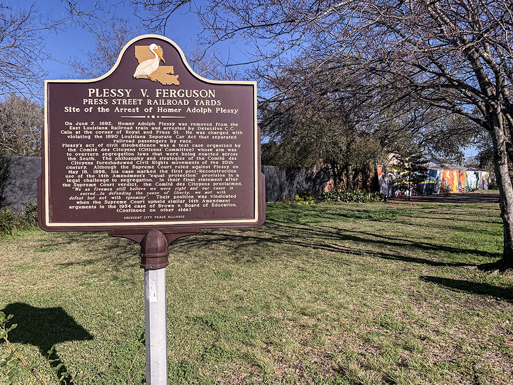 A historical marker for Plessy v. Ferguson at Press Street Railroad Yards, detailing the arrest of Homer Adolph Plessy. The sign is surrounded by trees and a pathway, with a colorful mural visible in the background.