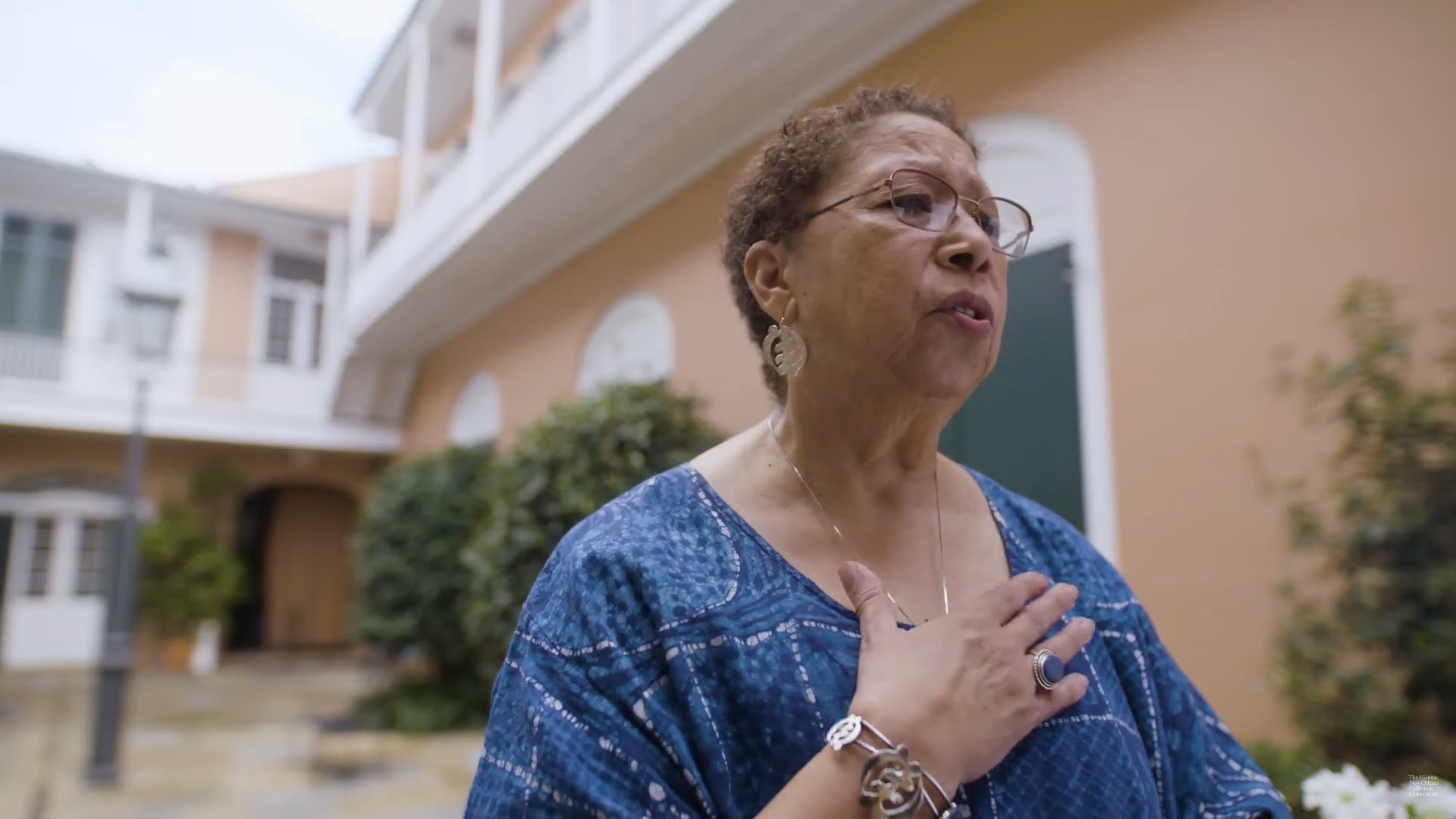 An older woman with short curly hair and glasses stands outdoors, wearing a blue top and earrings. She places her hand on her chest. The background features a peach-colored building with white trim and greenery.