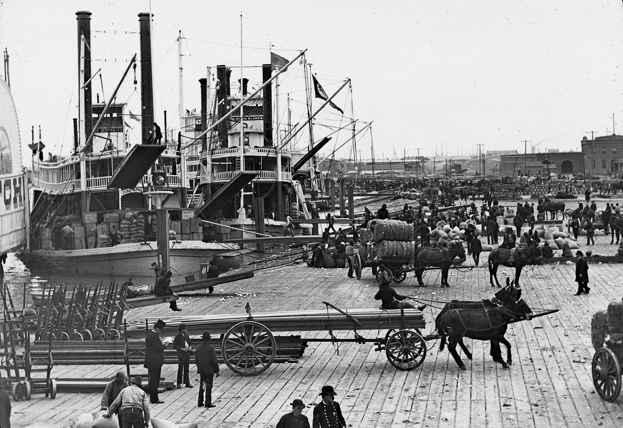 A historical black-and-white photo of a busy river dock with three large steamships. People and horses are unloading cargo, including barrels and bags, onto wooden carts. The scene is bustling with activity.