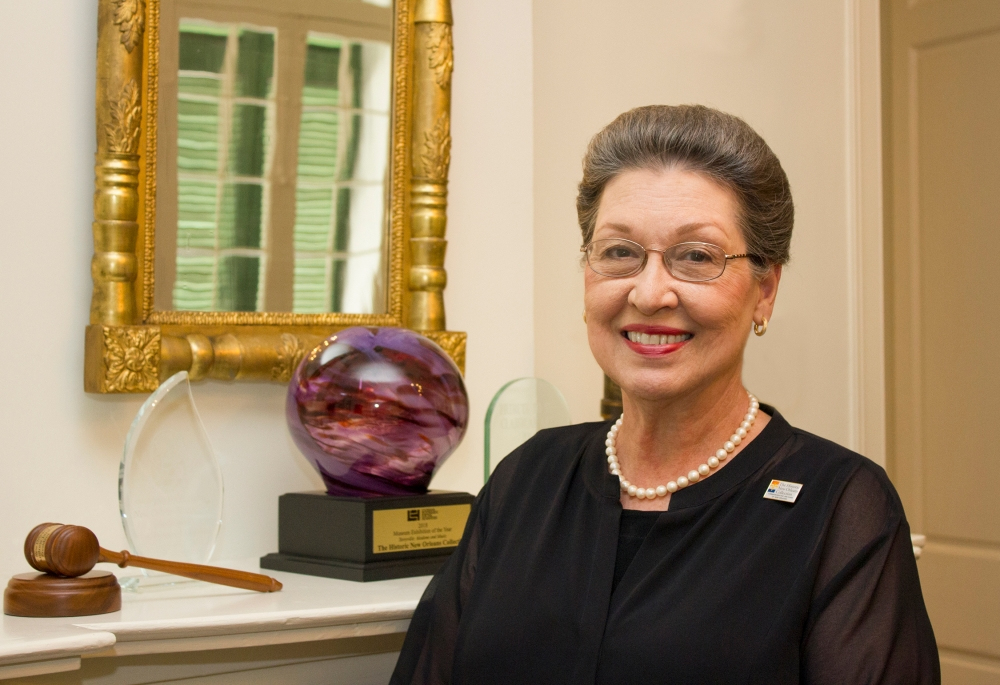 A woman with glasses and a pearl necklace smiles in front of a mirror. She is standing near a shelf with a gavel, glass awards, and a colorful glass orb. The background features a section of a window with green shutters.