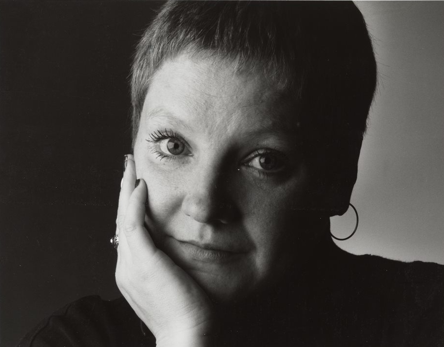 Black and white portrait of Leigh Harris with short hair, resting their head on their hand. They are looking directly at the camera, wearing a dark top and a hoop earring. The background is split into light and dark sections.