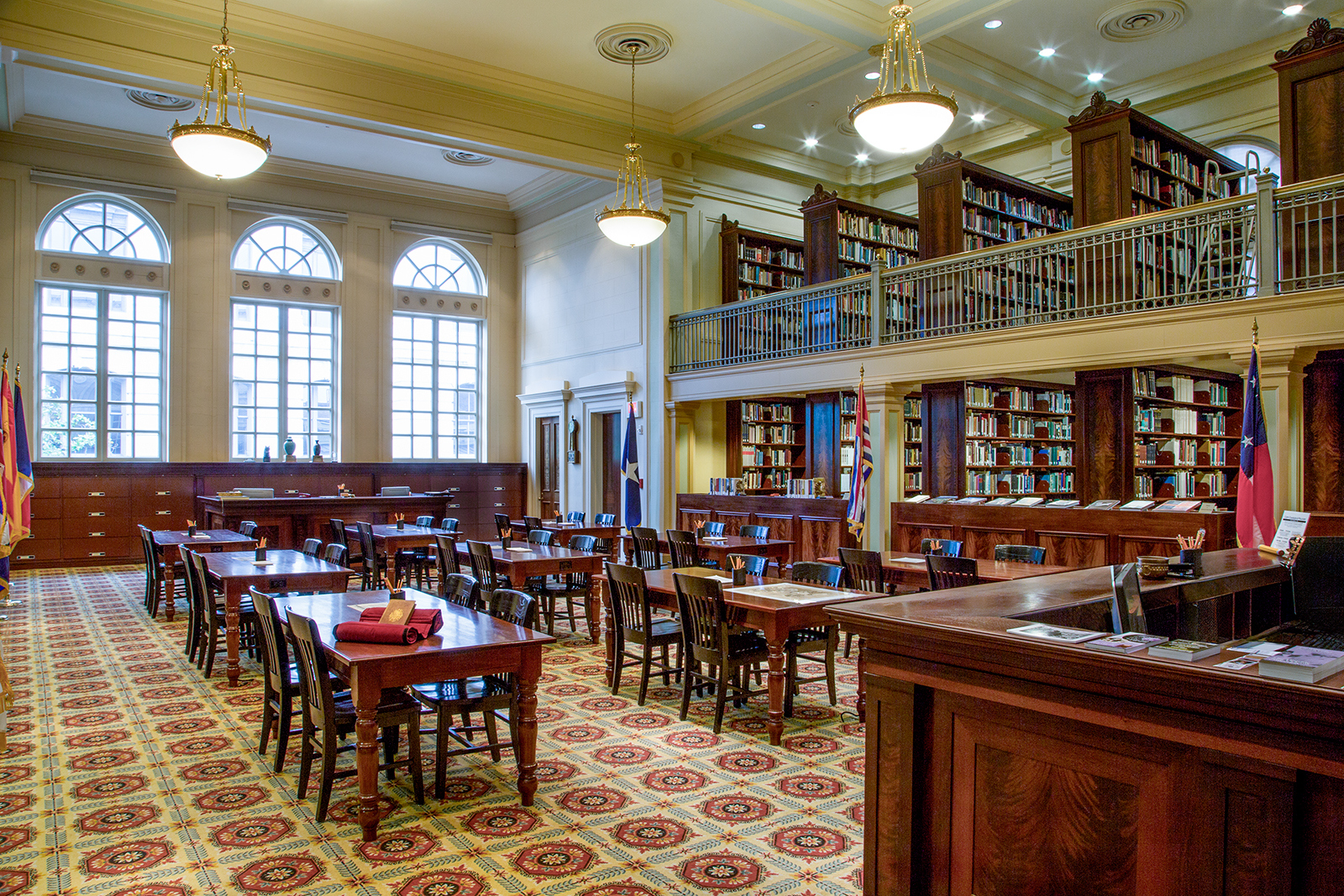 A spacious, elegant library with wooden tables and chairs arranged in rows. Bookshelves line the walls, extending to a second-floor balcony. Large arched windows and hanging lamps illuminate the room, highlighting the patterned carpet.