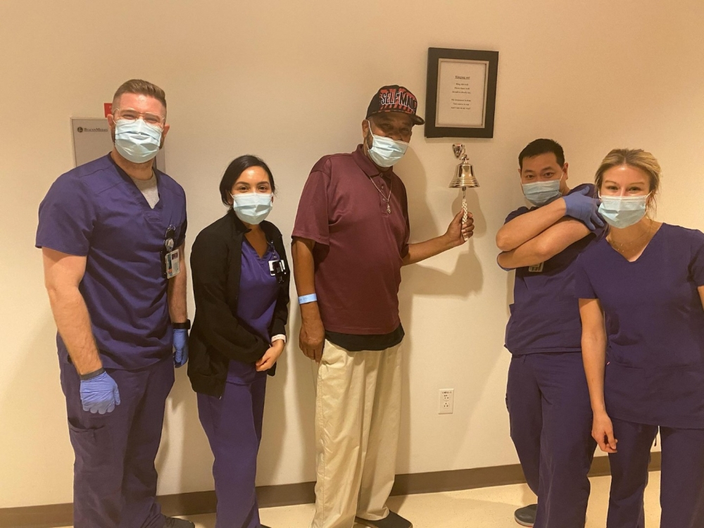 A group of healthcare workers in scrubs and masks stands with a man holding a bell on a stick. Everyone appears cheerful. The setting looks like a medical facility.