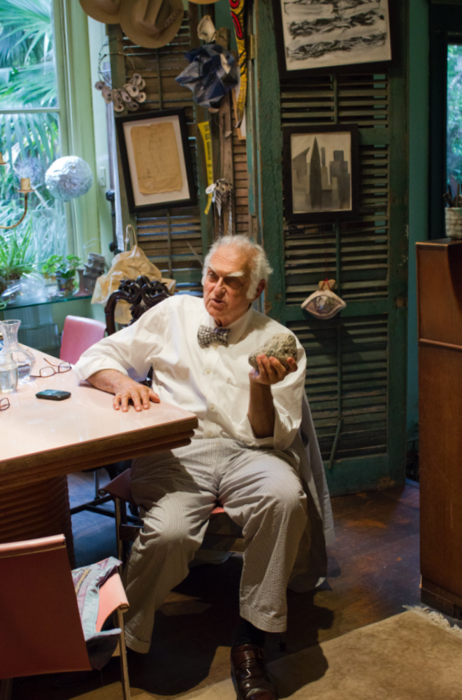 Elderly man wearing a bow tie sits at a table, holding a rock and gesturing. The room is filled with eclectic decor, including artwork and hats. Sunlight streams through a window with plants outside.