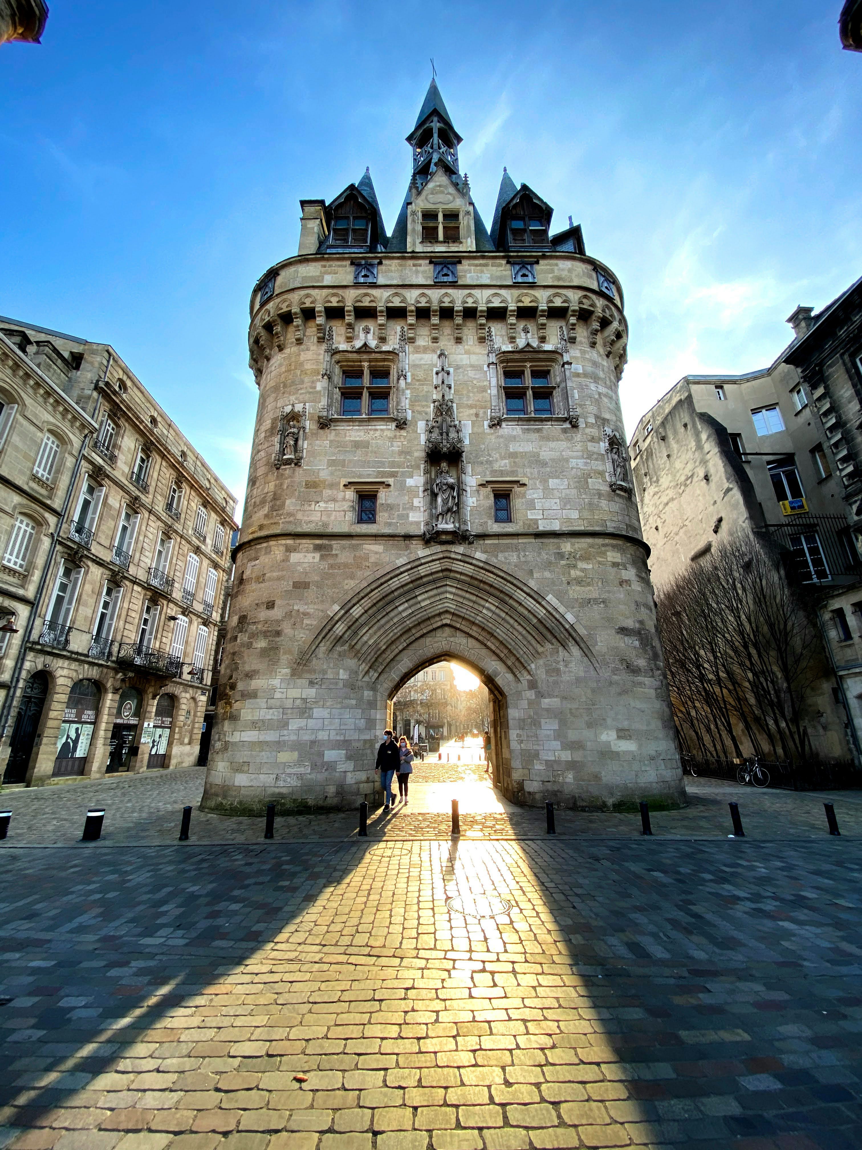 A historic stone gate with an arched passageway stands under a clear blue sky. Sunlight streams through, casting long shadows on the cobblestone path. Two people walk through the arch, surrounded by old buildings on either side.