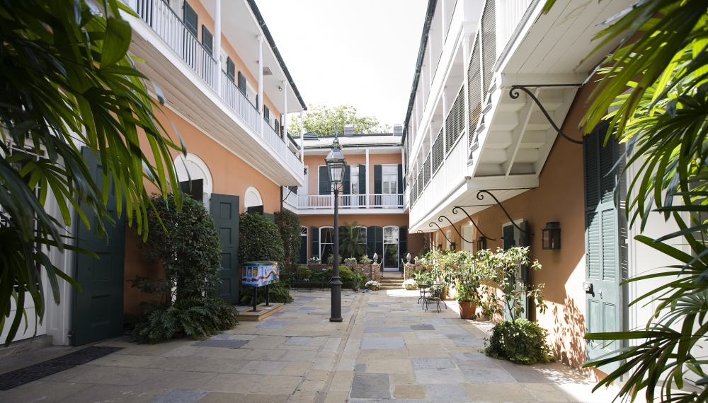 A charming courtyard with cobblestone pavement, surrounded by two-story buildings with balconies. There are potted plants, green shutters, and a street lamp in the center. Lush greenery frames the scene.