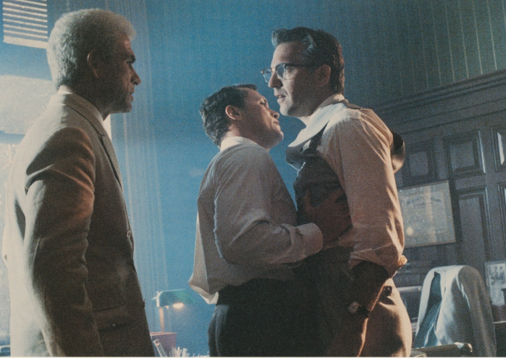 Three men in a dimly lit office. Two stand close, one grabs the others collar, appearing tense. The third man, with gray hair, observes. The room has wood paneling and a green desk lamp.
