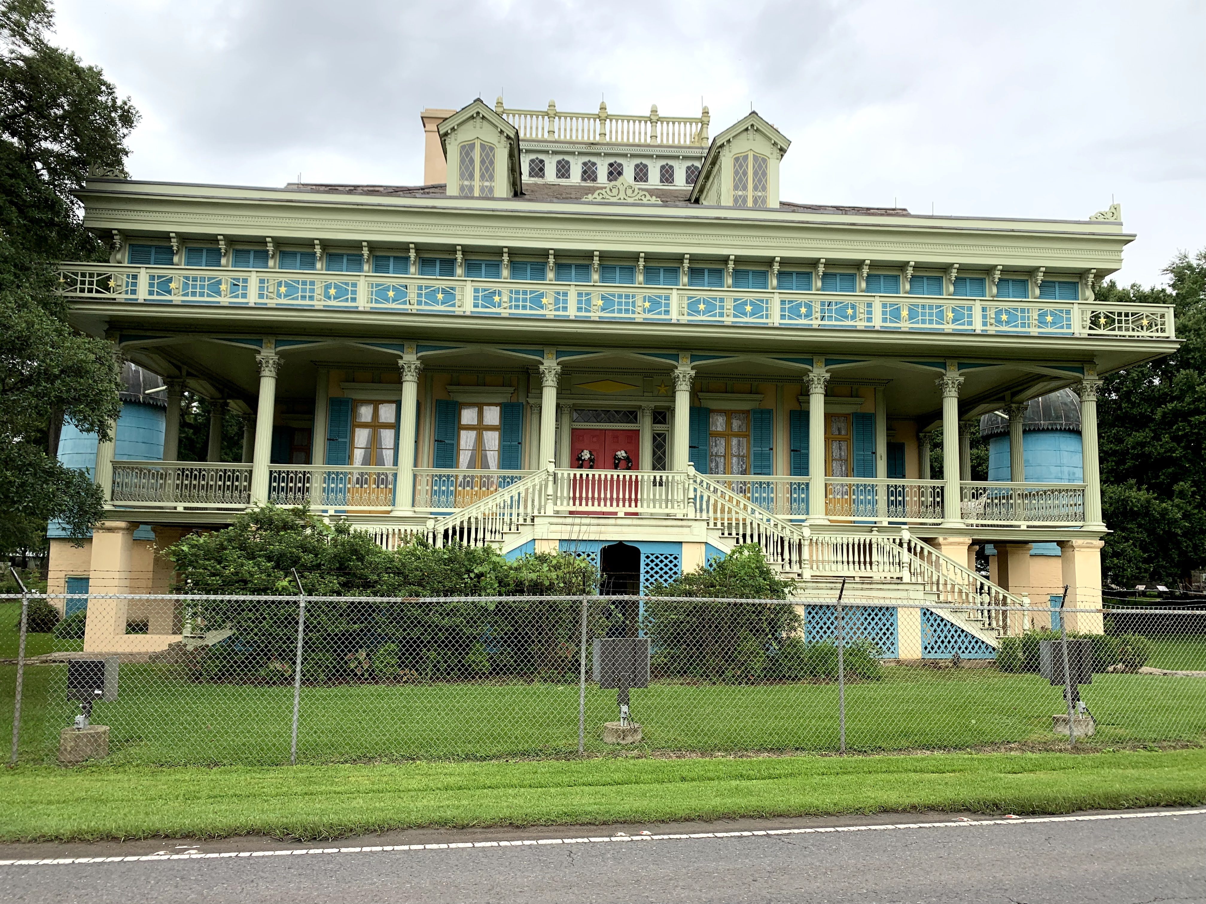A historic mansion with a vibrant turquoise and lime-green exterior, featuring a large porch with ornate columns and railings. The building is elevated and surrounded by greenery, with a chain-link fence in the foreground.