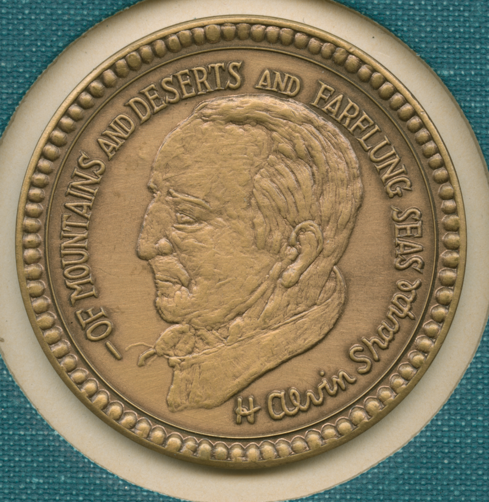 A bronze medallion featuring a profile of a man with the text Of Mountains and Deserts and Farflung Seas and the name H Alvin Sharp engraved on it. The medallion is set against a teal fabric background.