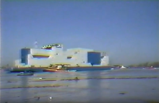 A screenshot from vintage footage of the Vernon C. Bain prison ship shows a  massive, hulking, boxy barge in the middle of a body of water, surrounded by smaller ships.