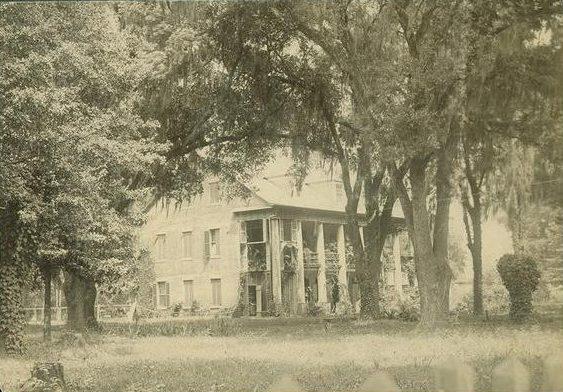 A sepia-toned image of a historic house with large pillars and a wraparound porch, surrounded by tall trees and a grassy lawn. The architecture suggests an old Southern plantation-style building set amidst a tranquil natural setting.