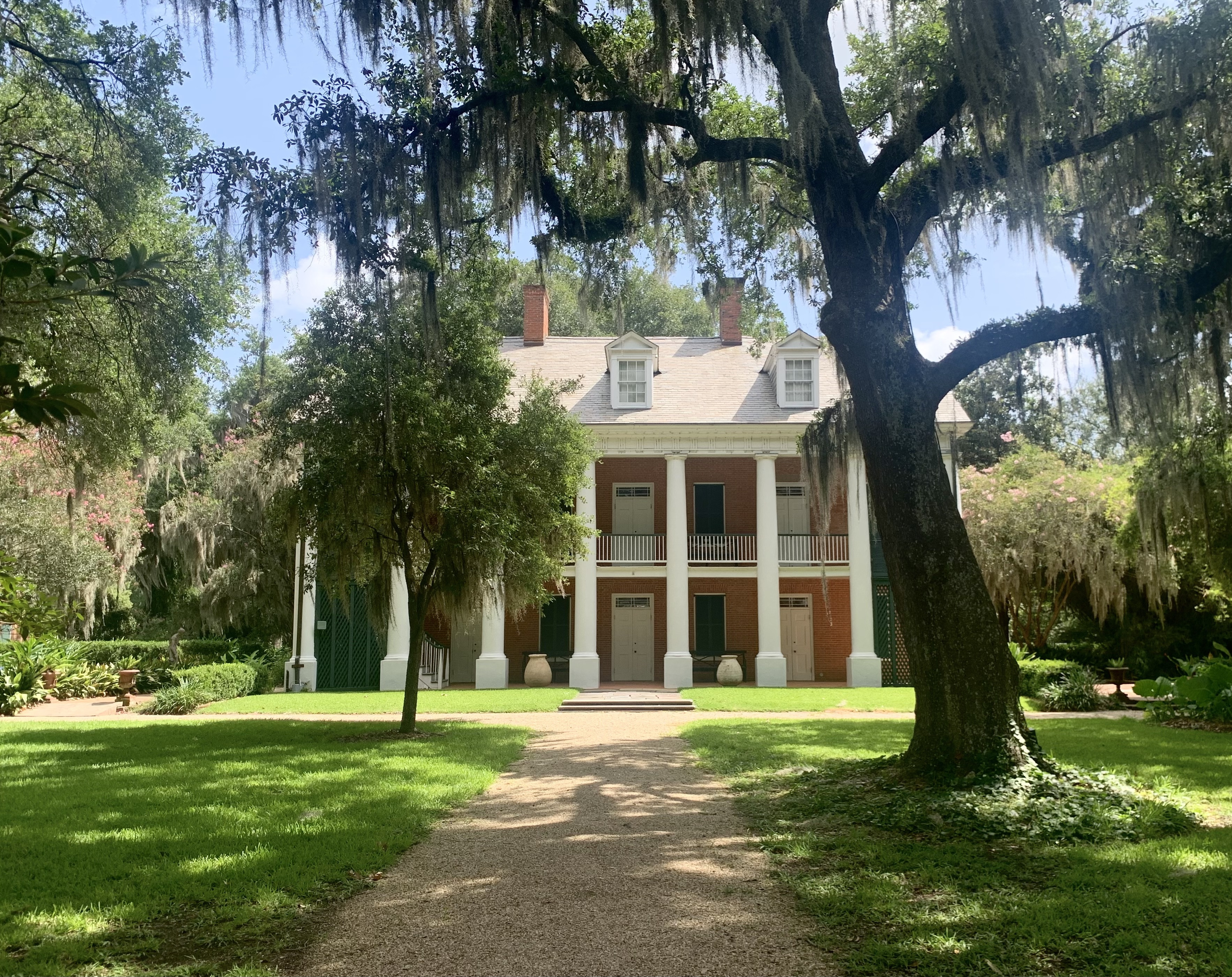 A two-story historic house with a white facade and brick accents is surrounded by lush greenery. A large oak tree with Spanish moss drapes over a pathway leading to the house. The scene is bathed in sunlight.