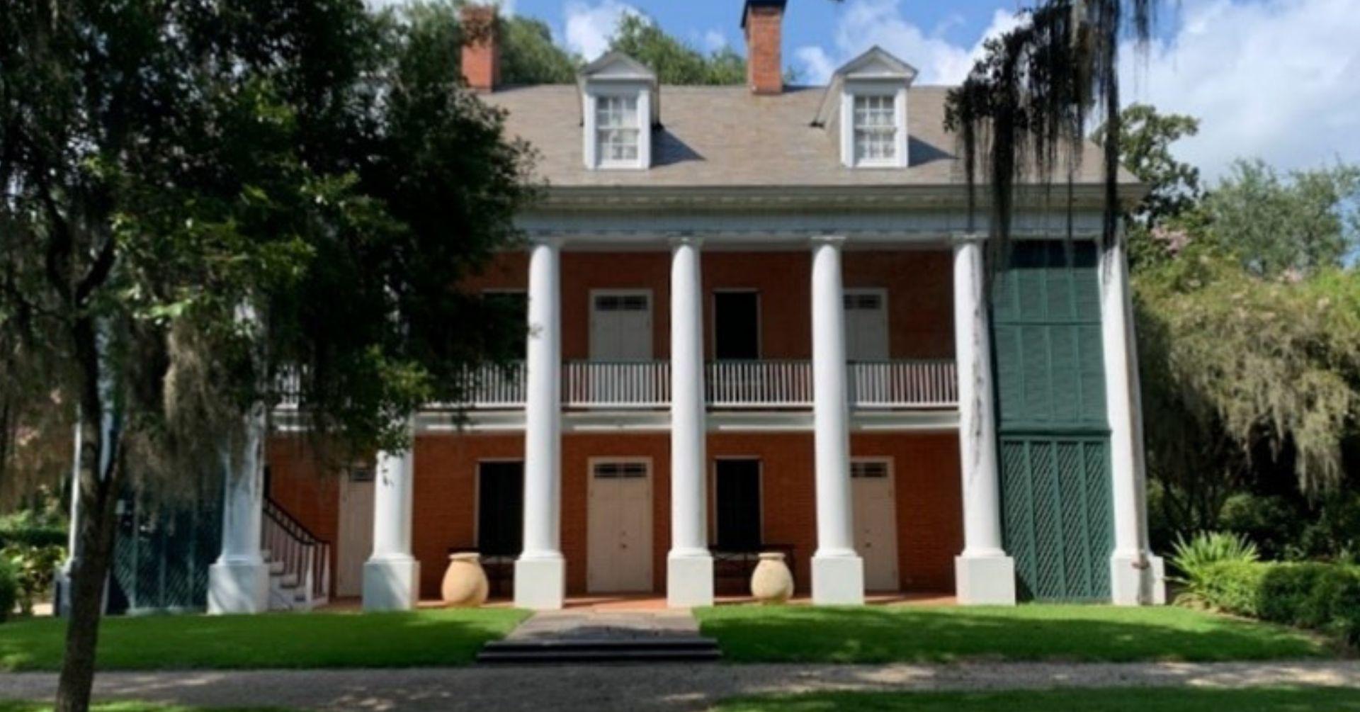 A historic two-story brick house with white columns and a balcony, surrounded by trees and green grass. Spanish moss hangs from the branches, and the sky is partly cloudy.