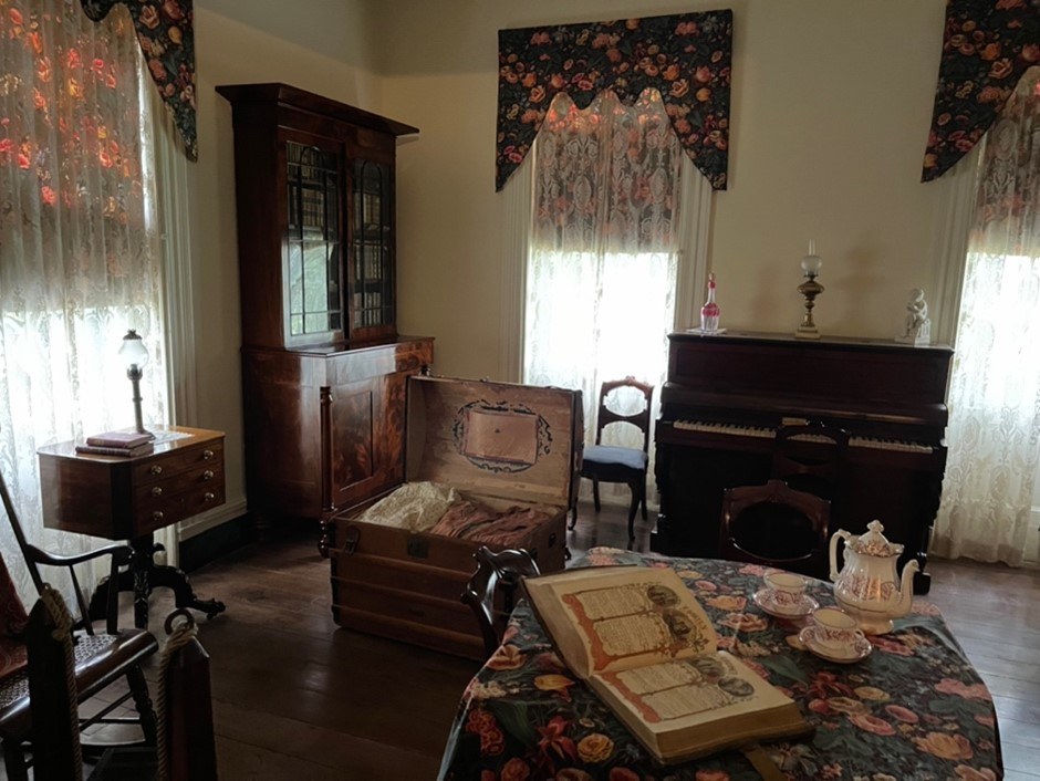 A vintage living room with a piano, ornate wooden furniture, and lace curtains. An open trunk sits on the floor, while a table is adorned with a teapot, teacups, and an open book. The room has a cozy, antique feel with floral accents.
