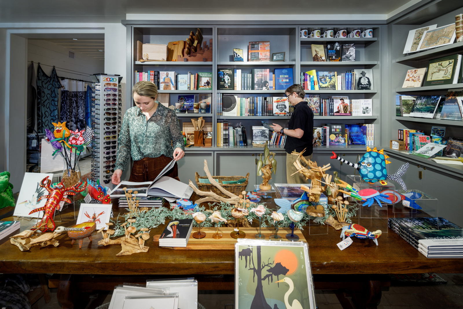 Two people browsing in a colorful bookstore filled with books, artwork, and decorative figures on display. One person flips through a book on a wooden table, while another examines books on a shelf in the background.