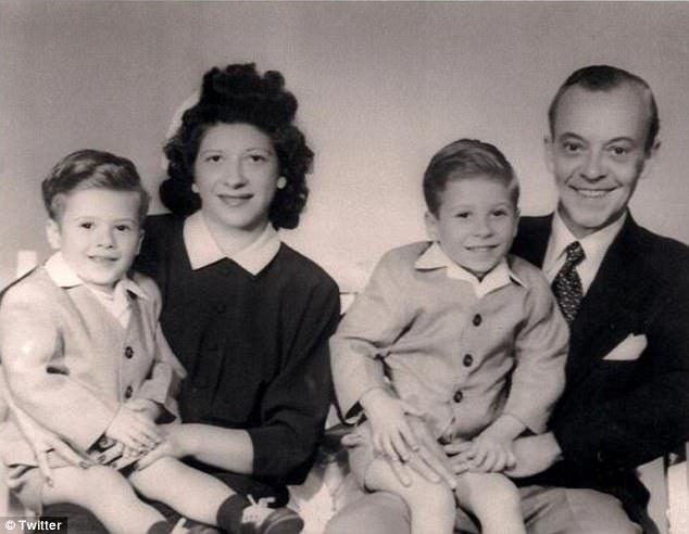 A vintage black and white photo of a family. Two young boys are sitting on the laps of a woman and a man. The woman and man are wearing formal attire. The boys are dressed in matching outfits with shorts and blazers.