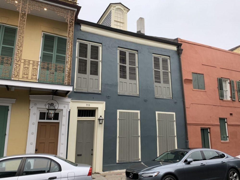 Street view of colorful historic buildings. The central building is painted gray with shutters on the windows, flanked by a yellow building with a balcony and a salmon-colored building. Cars are parked on the street in front.