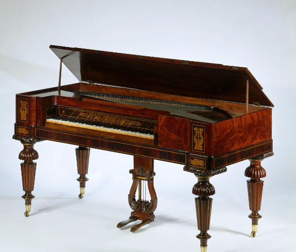 A vintage grand piano with a polished wood finish and intricate decorative details. The piano lid is open, displaying the strings and hammers inside. It stands on four ornate, turned legs with brass casters.