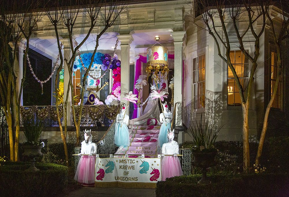 A decorated house features the Krewe of Unicorns theme. The porch has unicorn mannequins with colorful tutus and feathered headpieces. Bright purple, pink, and blue flowers adorn the entrance, creating a festive and whimsical atmosphere.