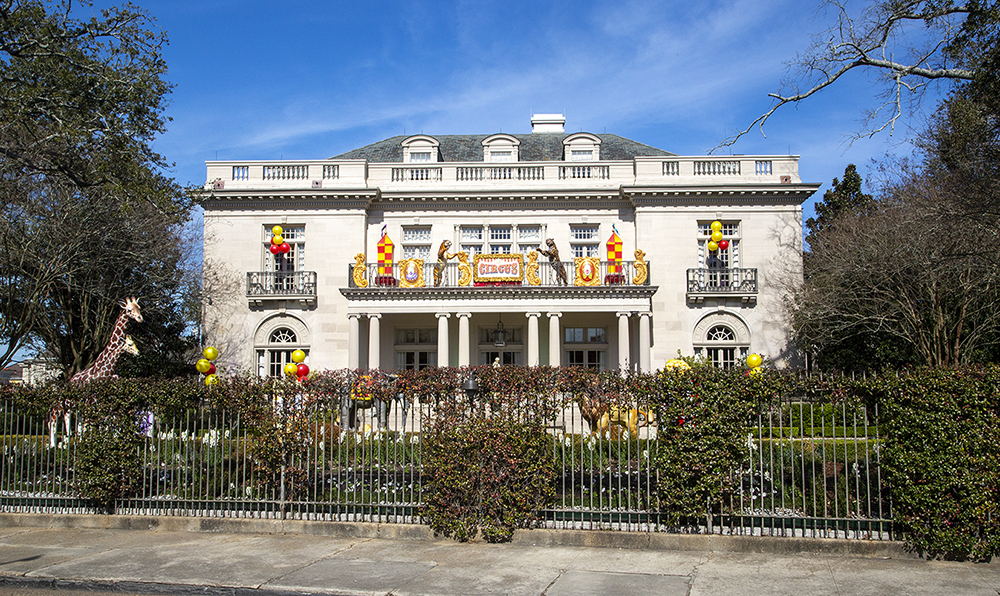 A grand white building with a decorative facade is adorned with colorful carnival-themed decorations. A giraffe sculpture stands on the left, and balloons are scattered around the garden, all under a clear blue sky.