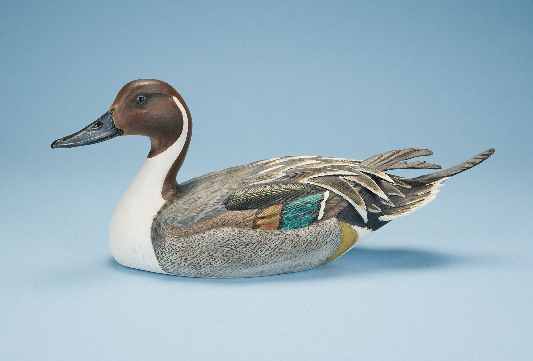 A detailed wooden carving of a duck against a light blue background, showcasing realistic textures and colors with a glossy brown head, white neck, and intricately painted wings in shades of gray, brown, and green.