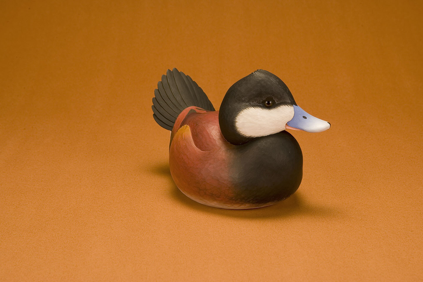 A detailed, intricately carved duck decoy with a blue bill, black head, and reddish-brown body is set against an orange background. Its tail feathers are raised, showcasing craftsmanship.