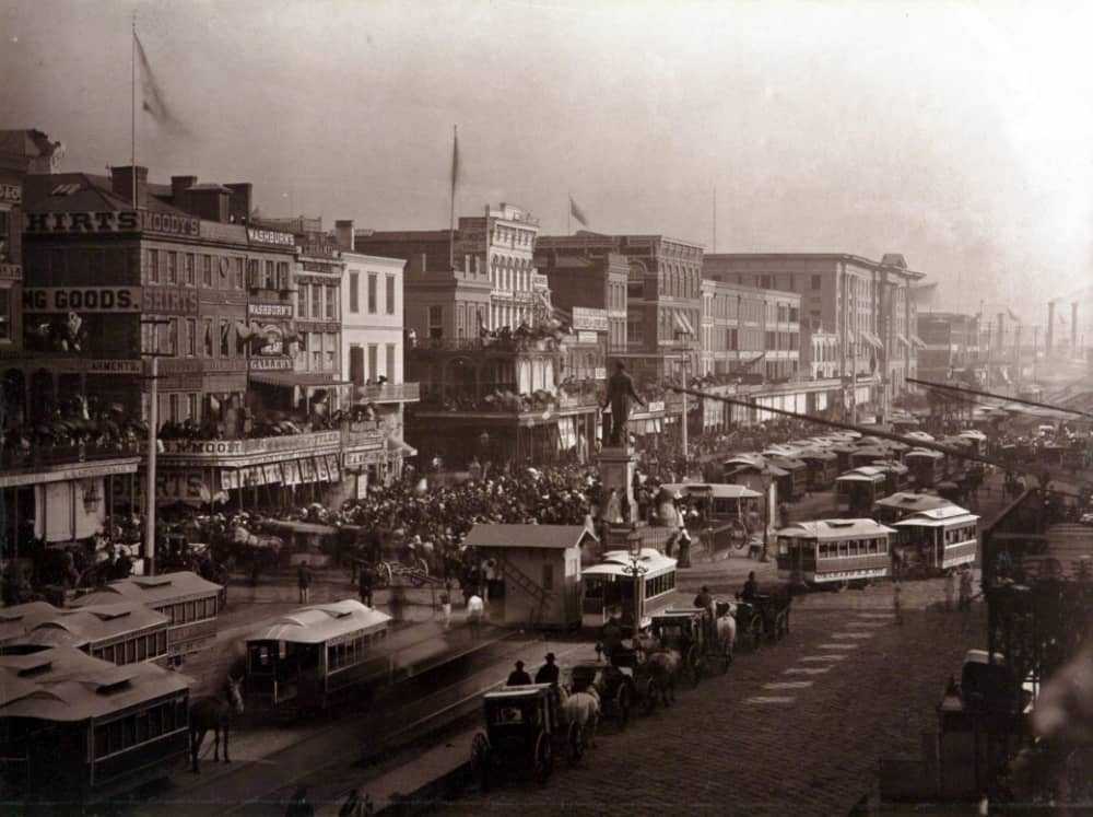 A bustling 19th-century street scene featuring horse-drawn carriages, streetcars, and busy pedestrians. Buildings adorned with advertisements line the street, adding to the vibrant urban atmosphere.