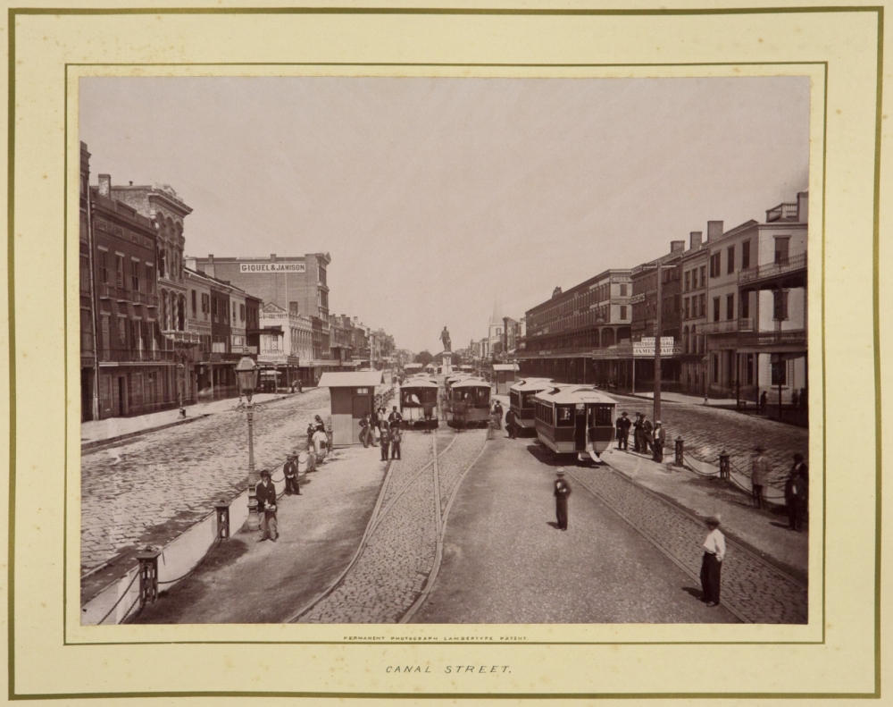 Black and white photo of Canal Street featuring vintage streetcars and people along the cobblestone road. Buildings line both sides of the street, and various pedestrians and horse-drawn carriages are visible.