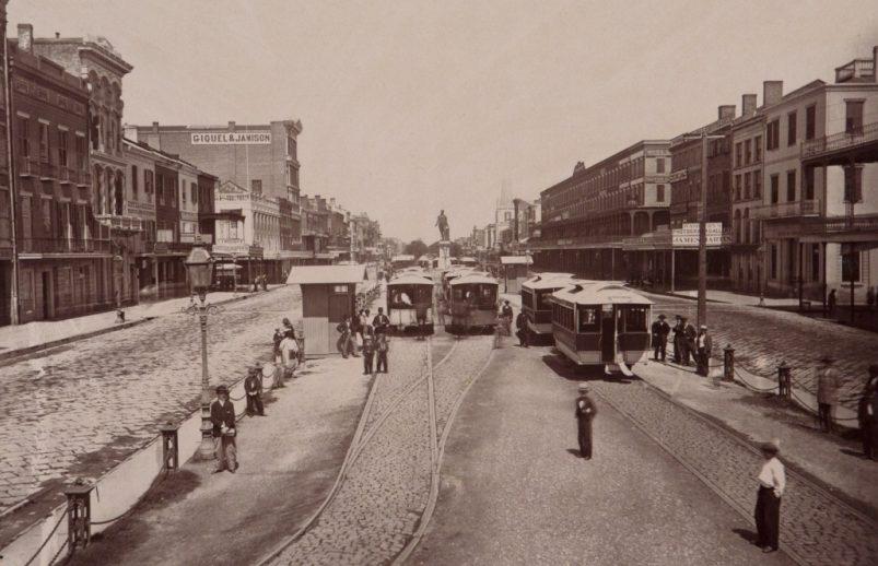 A black and white photograph of a historic city scene with cobblestone streets. Horse-drawn trams are on railroad tracks, and people are walking around. Buildings line the street, and there is a statue in the background.