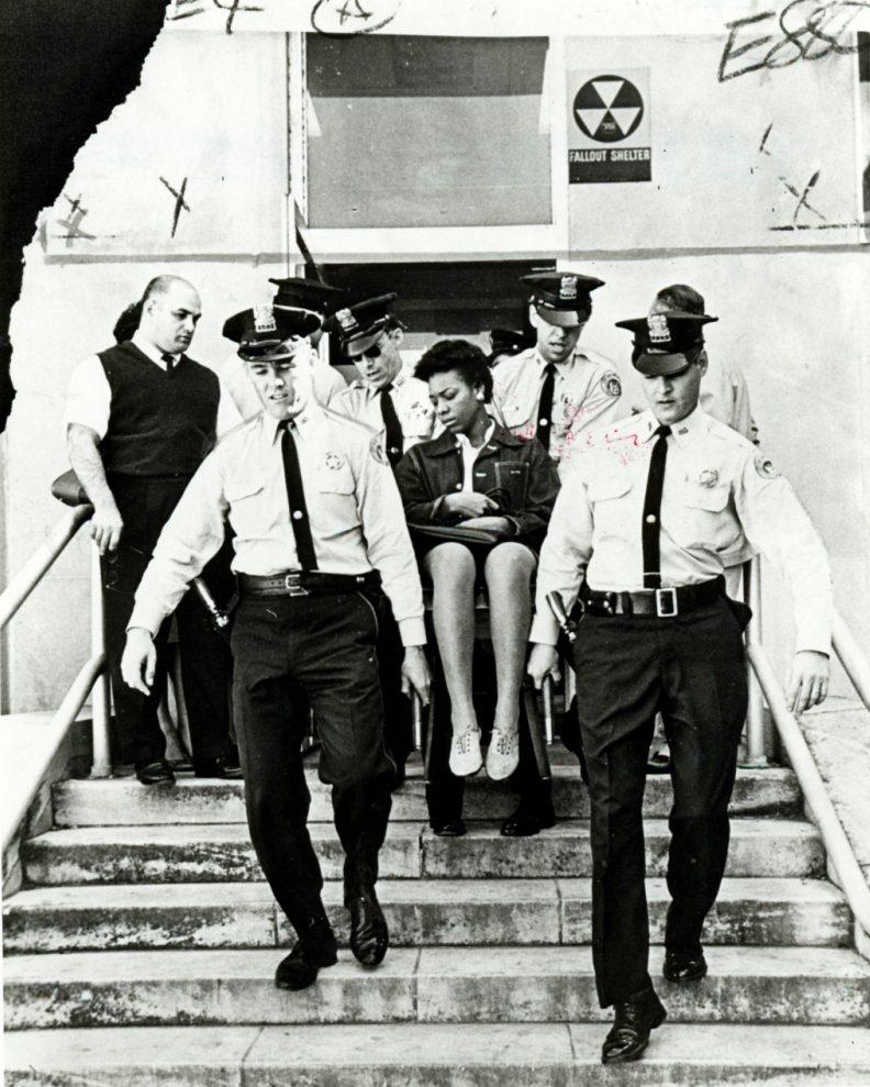 A group of police officers escort a seated woman down the steps of a building with a fallout shelter sign. The scene is black and white, and the officers are in uniform. The background includes a doorway and concrete steps.