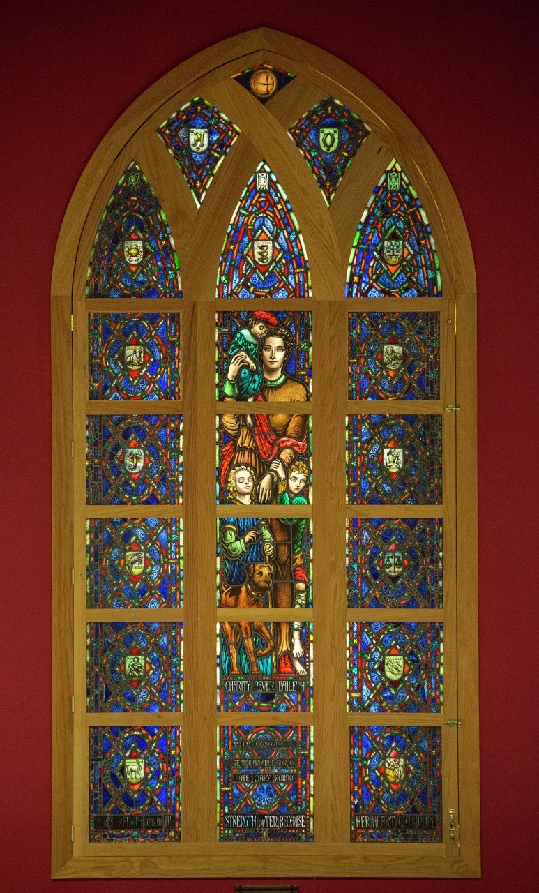 A colorful stained glass window with various intricate patterns and depictions of people, animals, and text panels. The narrow, arched design features sections with medieval style art against a rich red background.